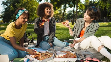 Drei fröhliche Frauen sitzen auf Wiese im Park auf einer Picknickdecke und essen Pizza. Frau links greift nach einem Stück Pizza, die anderen Frauen halten ein Stück in der Hand. Vor ihnen zwei Pizzakartons, Notizbücher, Kopfhörer, Bücher.