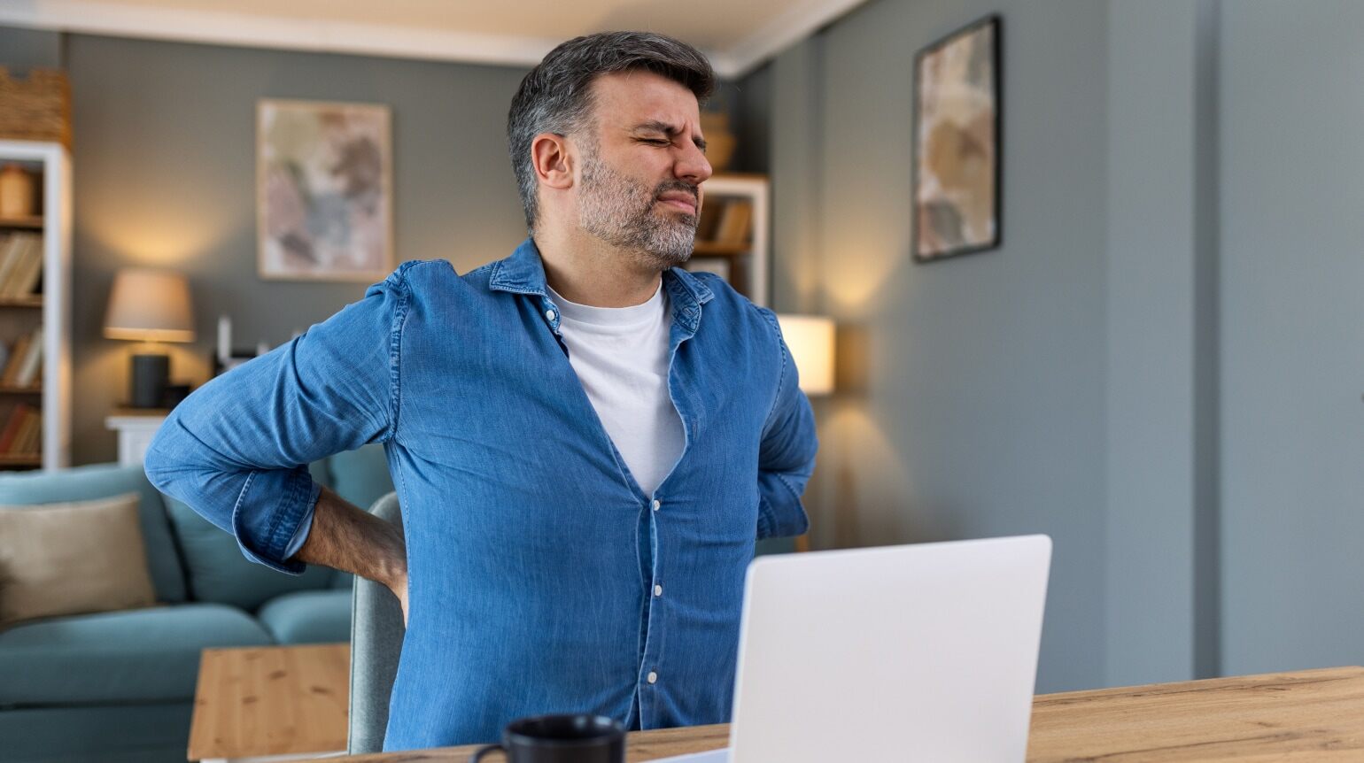 Homeoffice: Mann im weißen Shirt, hält sich den schmerzenden Rücken. Er sitzt vor einem Laptop auf Holztisch, links eine Tasse, im Hintergrund Sofa und Regal.