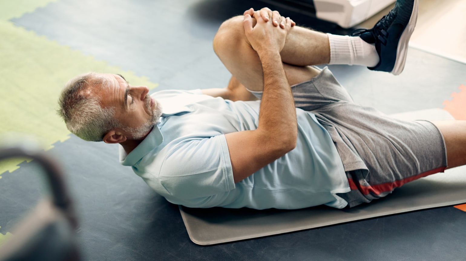 Seitenansicht von einem Mann in Sportkleidung. Er liegt auf dem Rücken auf einer Matte im Fitnessstudio und dehnt das linke Bein, indem er das Knie an seine Brust zieht. Das rechte Bein ist ausgestreckt. Im Hintergrund ein Laufband.