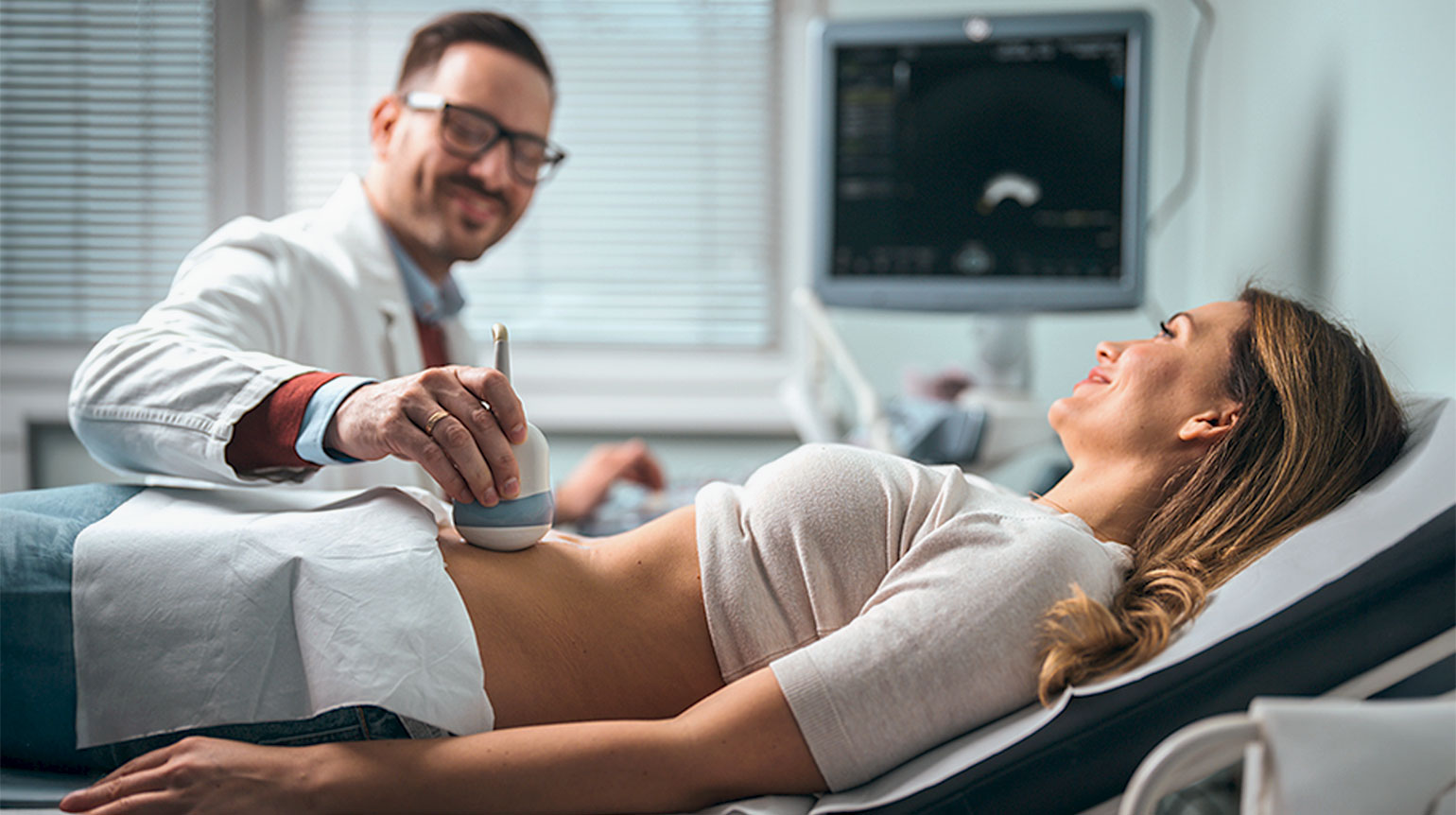 Arzt macht eine Ultraschalluntersuchung des Bauchs bei Patientin. Beide lächeln. Sie liegt auf einer Liege und trägt ein helles, nach oben gezogenes Oberteil. Ihr Unterleib ist bedeckt. Im Hintergrund das Ultraschallbild auf einem Monitor.