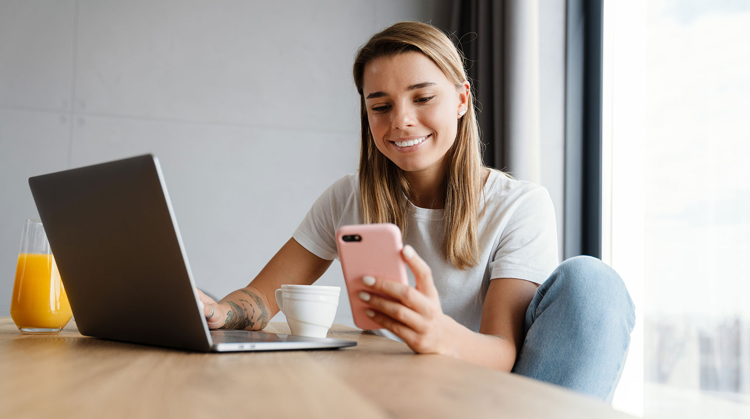 Frau sitzt an einem Holztisch, vor sich ein aufgeklapptes Laptop, eine Tasse und ein Glas Orangensaft. Sie hält in der linken Hand ein rosafarbenes Mobiltelefon und schaut lächelnd darauf. Die rechte Hand liegt auf der Tastatur des Laptops.