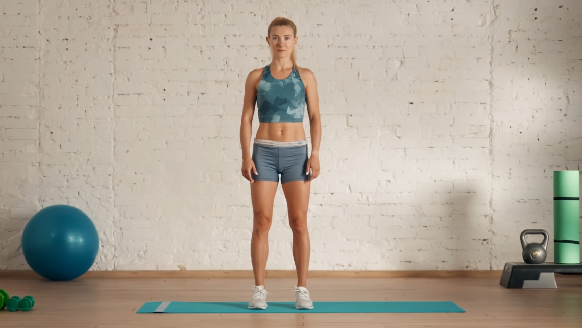 Vorderansicht von einer Frau in Sportkleidung. Sie steht aufrecht, Füße hüftbreit auseinander, in einem Raum mit einem blauen Sitzball, einer Gymnastikmatte, einem Steppbrett, einem Kettle Dumbbell und einer Faszienrolle.