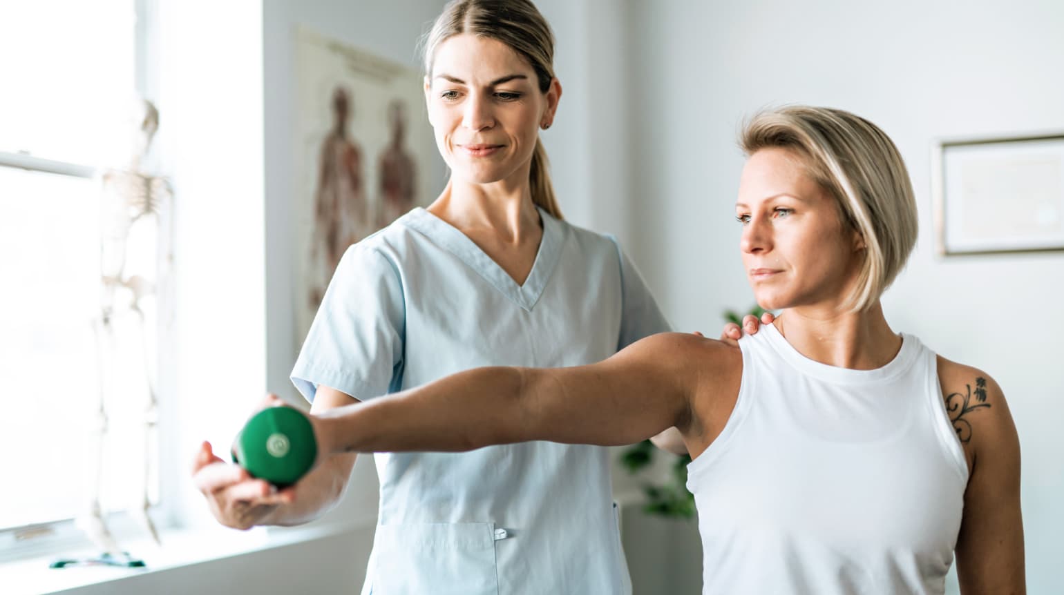 Vorderansicht von zwei Frauen, links Physiotherapeutin, rechts Patientin. Die Patientin streckt ihren rechten Arm aus, hält eine kleine grüne Hantel in der Hand. Die Therapeutin korrigiert ihre Haltung. Im Hintergrund ein Behandlungszimmer mit Anatomieposter und Fenster.