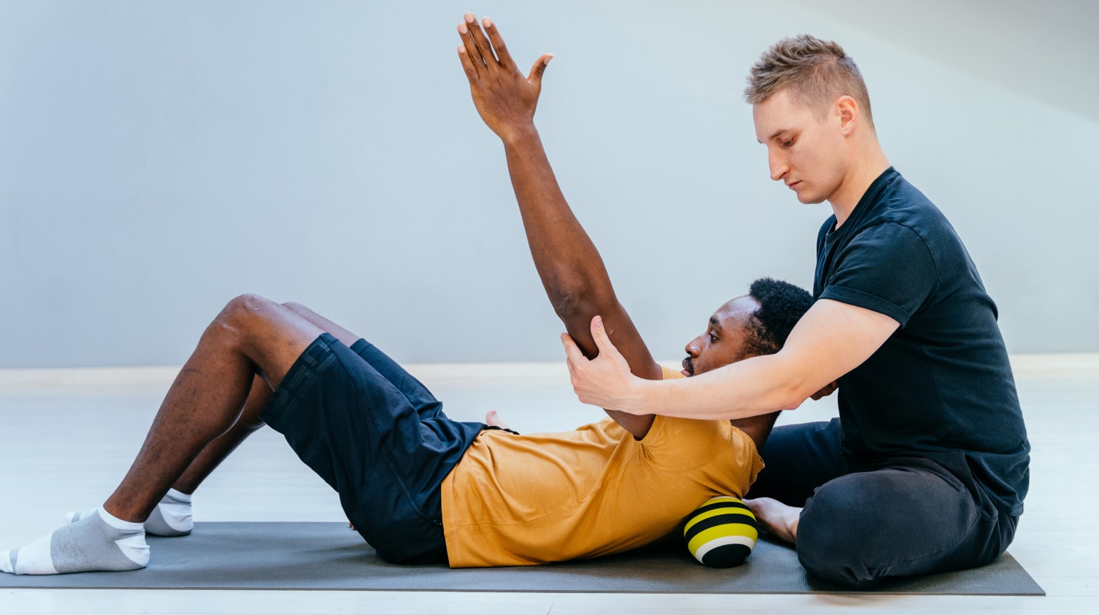 Schulterübung: Zwei Männer in Sportkleidung auf einer Sportmatte. Ein Mann liegt auf dem Rücken mit aufgestellten Beinen und hebt beide Arme ausgestreckt schräg nach oben. Sein Oberkörper liegt auf einer Rolle. Der zweite Mann sitzt hinter seinem Kopf und stabilisiert die Armposition.