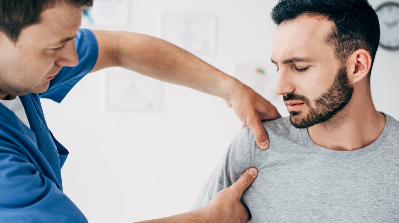 Ein blau gekleideter Therapeut/Arzt in der Seitenansicht umfasst mit beiden Händen das Schultergelenk des Patienten rechts von ihm. Der Patient in der Frontalansicht trägt ein graues Shirt und blickt auf die Hände des Therapeuten/Arztes.